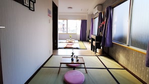 Una habitación tradicional japonesa con suelo de tatami, futón, una pequeña mesa con un jarrón y una puerta corredera con cortinas moradas.