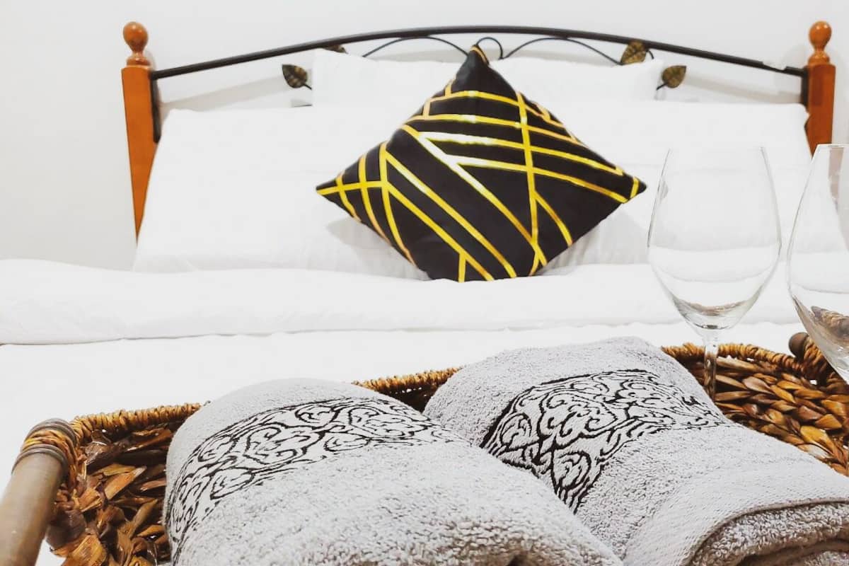Een net opgemaakt bed met een zwart-geel kussen, twee glaasjes water en een gevlochten mand met handdoeken.