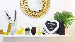 A white shelf with decorative items including a golden sunburst mirror, a heart-shaped sign with "WELCOME TO BALAI AMIGO," a potted plant, and various figurines.