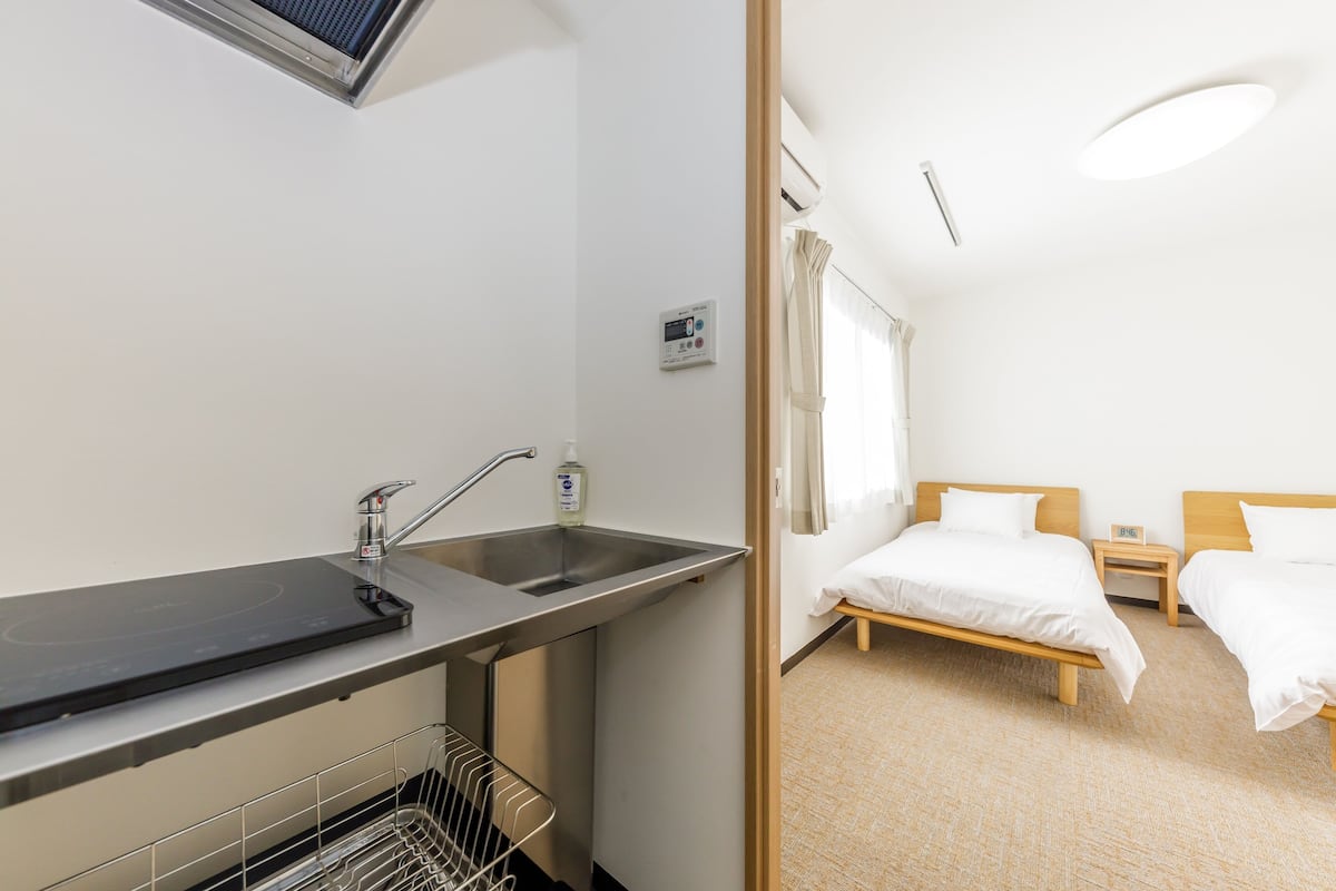 A hotel room with a kitchenette, a bed, a bedside table, and a window with curtains.