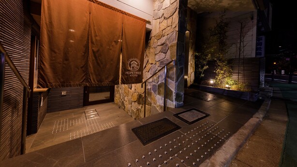 A hotel entrance with a stone wall, a brown curtain, and a sign that reads "HOPETREE KYOTO".