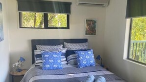 Een slaapkamer met een bed, twee ramen met zonwering, een airconditioningapparaat, een klein tafeltje met een lamp en een schilderij aan de muur.
