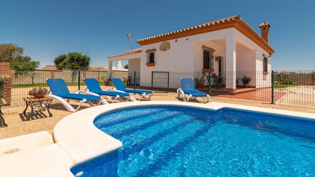 A swimming pool with lounge chairs, a white building with a red roof, and a clear blue sky. - Charming Home with Pool, Spacious Terrace, Garden & Wi-Fi; Pets Allowed, Parking Available (Conil de la Frontera)