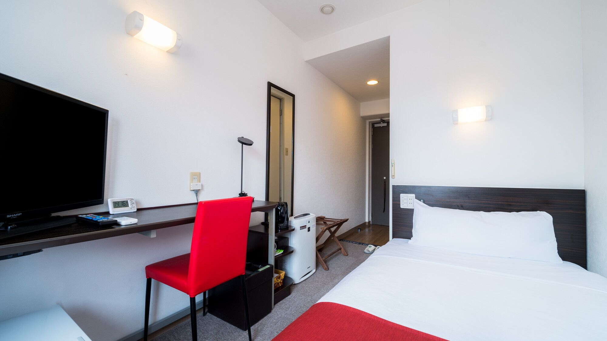 Ein Hotelzimmer mit Bett, Schreibtisch mit Fernseher, rotem Sessel, Spiegel und Kaffeemaschine.