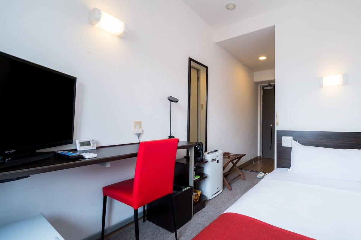A hotel room with a bed, a desk with a TV, a red chair, a mirror, and a coffee machine.