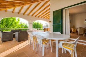 Un patio al aire libre con una mesa redonda, sillas blancas, mobiliario de mimbre y vistas a la vegetación.