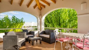 Een overdekt terras met rieten meubilair, een ronde tafel en uitzicht op groen en gebouwen.
