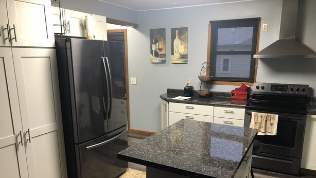 A modern kitchen with white cabinets, a black refrigerator, a granite countertop, and a stainless steel range hood. - The Farmhouse...only in town! (Sergeant Bluff)