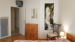 Une chambre avec une commode en bois, une chaise, un lit recouvert d’une courtepointe, un téléviseur fixé au mur et un tableau représentant une cascade.