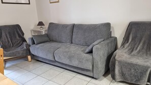 A grey sofa with matching armchairs, a small side table with a lamp, and a tiled floor.