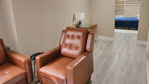 A hotel room with a massage chair, a small table with a lamp, and a bed with a blue bedspread.