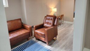 A room with a brown leather sofa, a matching armchair, a small table with a lamp, and a dining area in the background.