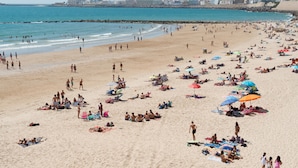 Ein überfüllter Strand mit Menschen, die sich sonnen und schwimmen, einem steinernen Wellenbrecher und Gebäuden im Hintergrund.
