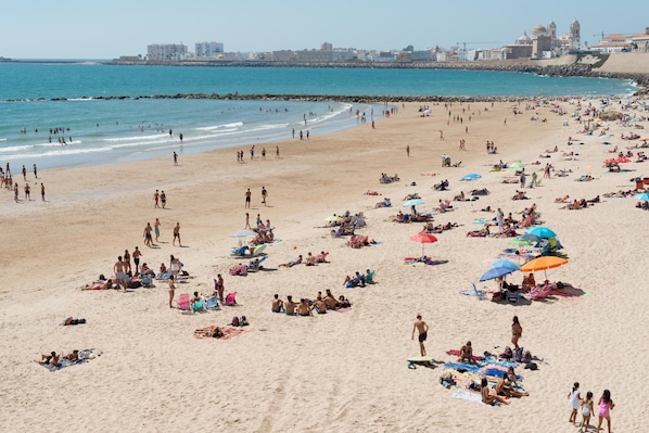 A crowded beach with people sunbathing and swimming, a rocky breakwater, and buildings in the background.