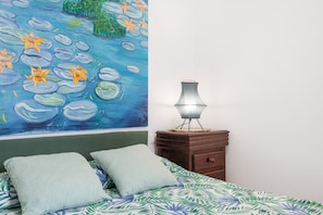 A bedroom with a bed, pillows, a nightstand, a lamp, and a wall painting.