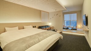 Ein modernes Hotelzimmer mit einem großen Bett, einer kleinen Sitzecke mit Tisch, einem an der Wand montierten Fernseher und einem großen Fenster mit Blick.