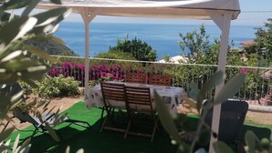 An outdoor patio with a table and chairs, surrounded by greenery and a view of the sea. - Apartment 'Il Rosmarino' with Sea View, Wi-Fi and Air Conditioning (Patresi)