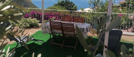 An outdoor patio with a table and chairs, surrounded by greenery and a view of the sea.