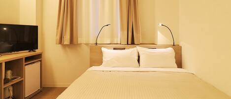 Ein Hotelzimmer mit Bett, Fernseher auf einem Holzboden, Schreibtisch mit Lampe und einem Fenster mit Vorhängen.