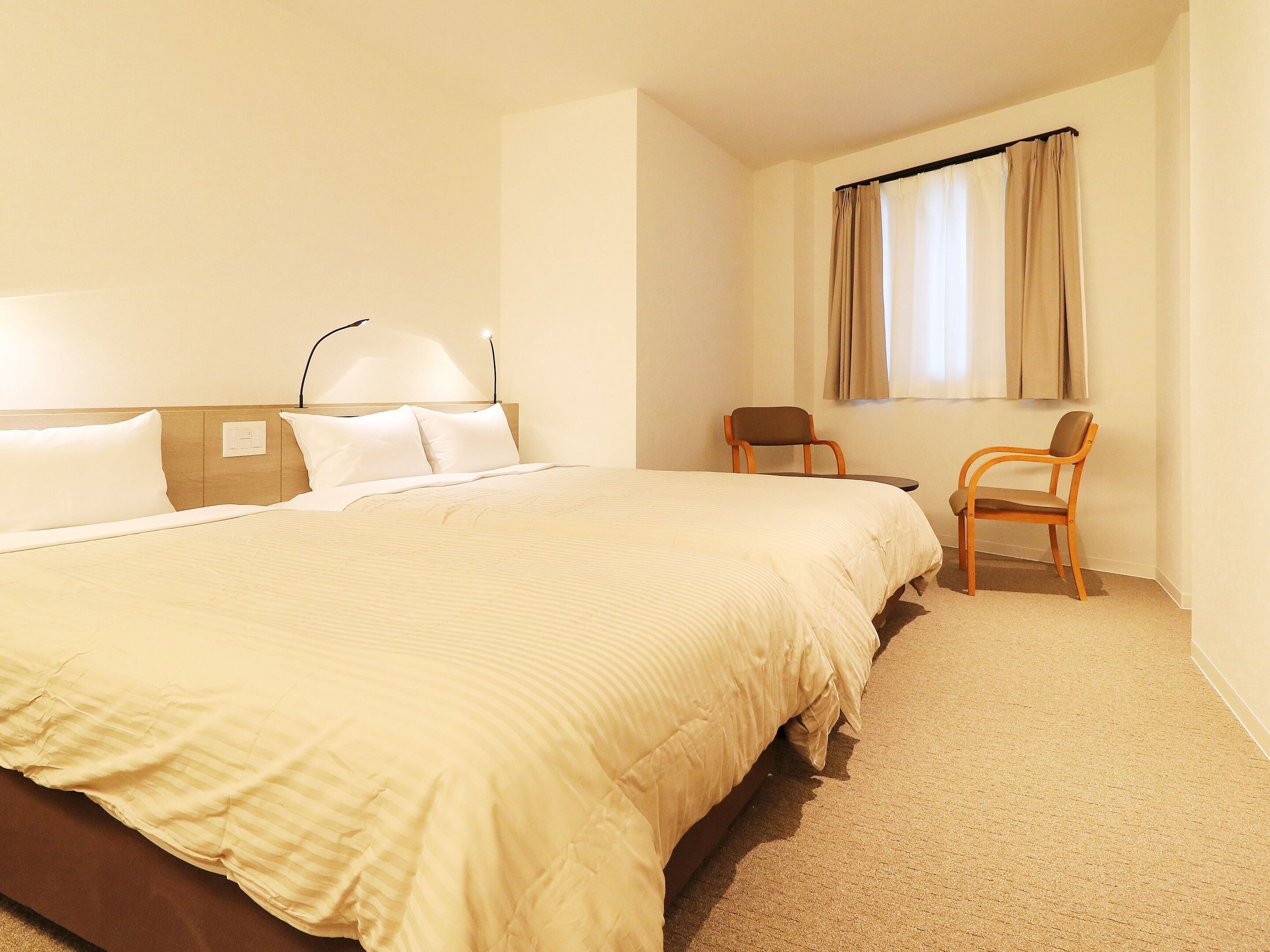 Habitación de hotel con una cama grande, dos sillas, una ventana con cortinas y una lámpara fijada a la pared.