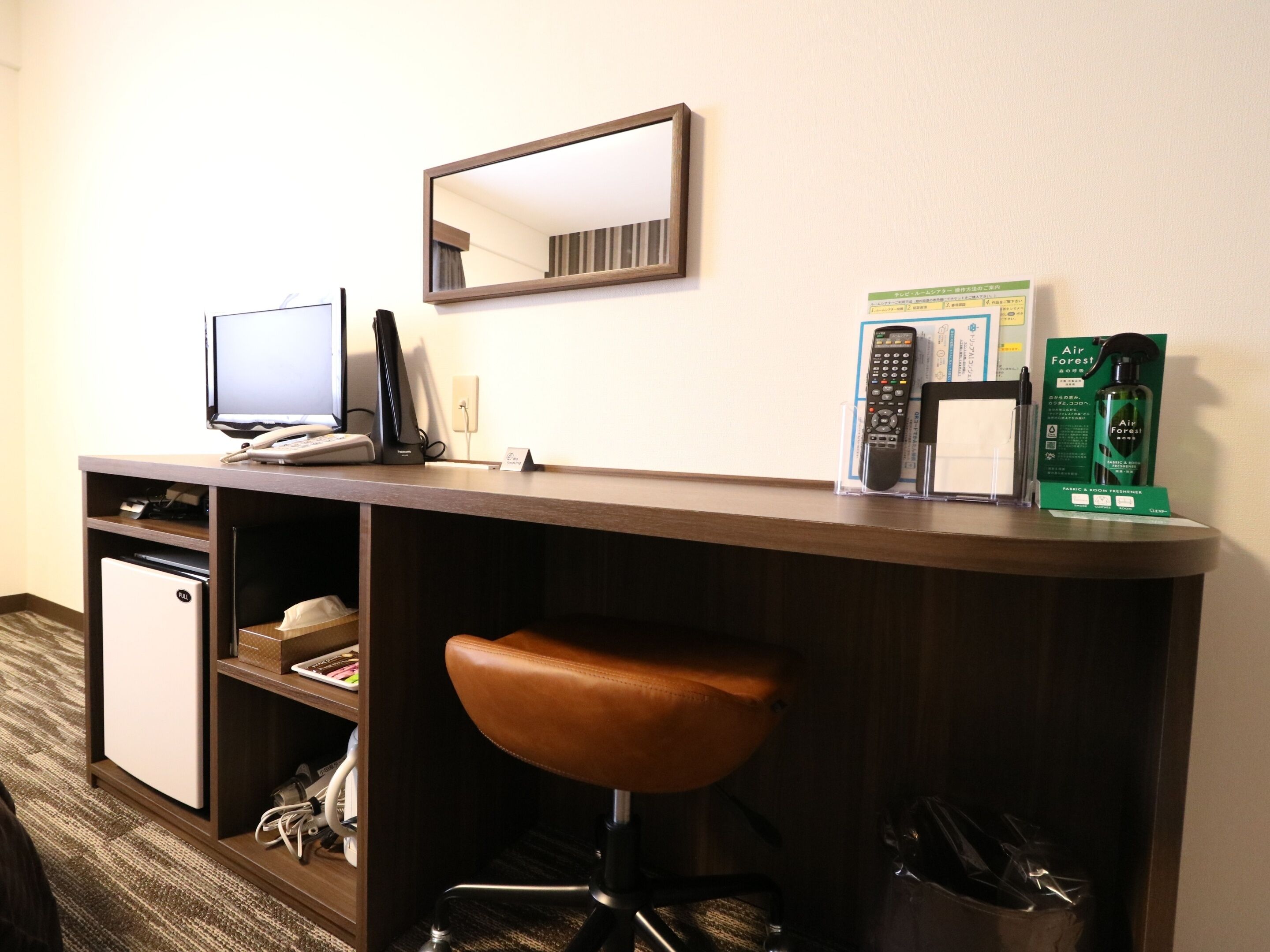 Habitación de hotel con escritorio, una silla, un refrigerador pequeño, un espejo, teléfono, control remoto y un ambientador Air Forest.