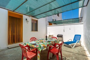 Une terrasse couverte avec une table dressée pour le repas, des chaises rouges, un fauteuil bleu et une fenêtre avec des rideaux.