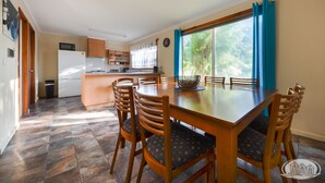 A dining area with a wooden table and chairs, a kitchen in the background, and a view of greenery through a window. - A quiet retreat just a stroll from the ocean (Allestree)