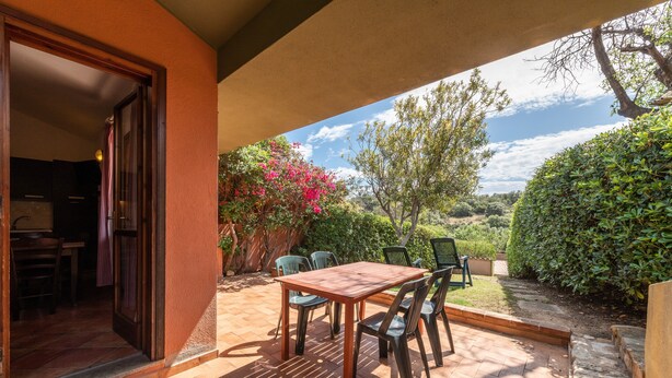 An outdoor patio with a wooden table and chairs, a covered seating area, and a view of a garden with various plants and trees. - Holiday home on the sea, immersed in nature – Accu is Prezzus E special (Villasimius)