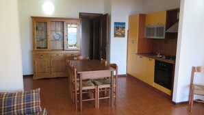 A compact living space with a dining table, chairs, a sofa, a kitchen area with cabinets and appliances, and a door leading to another room. - Sea View House Near the Beach – Casa Accu is Prezzus F (Villasimius)