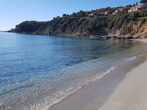 Ein Küstenstrand mit klarem Wasser, einem Sandstrand und Gebäuden an einem Hügelhang.