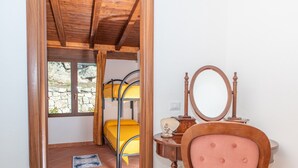 A room with a wooden ceiling, a bed with a yellow mattress, a wooden chair with a mirror, and a stone wall.