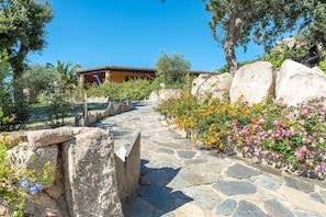 A stone pathway leading to a building with a tiled roof, surrounded by large rocks and flowering plants.