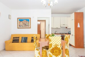 A living room with a sofa, a dining table, and chairs, a kitchen in the background, and a painting on the wall.