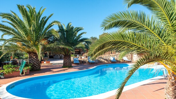 A swimming pool with palm trees, lounge chairs, and a stone building in the background.