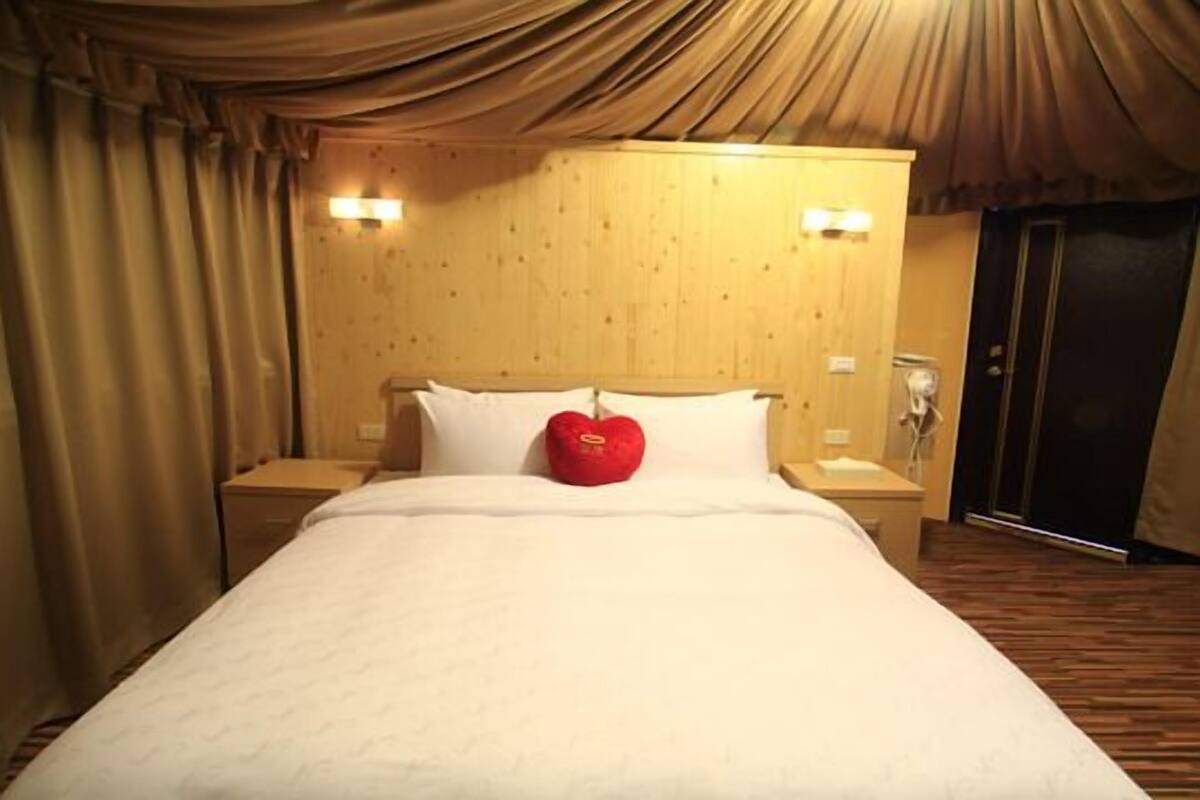 Habitación de hotel con una cama grande, cabecera de madera, dos lámparas de noche y un cojín rojo con forma de corazón.