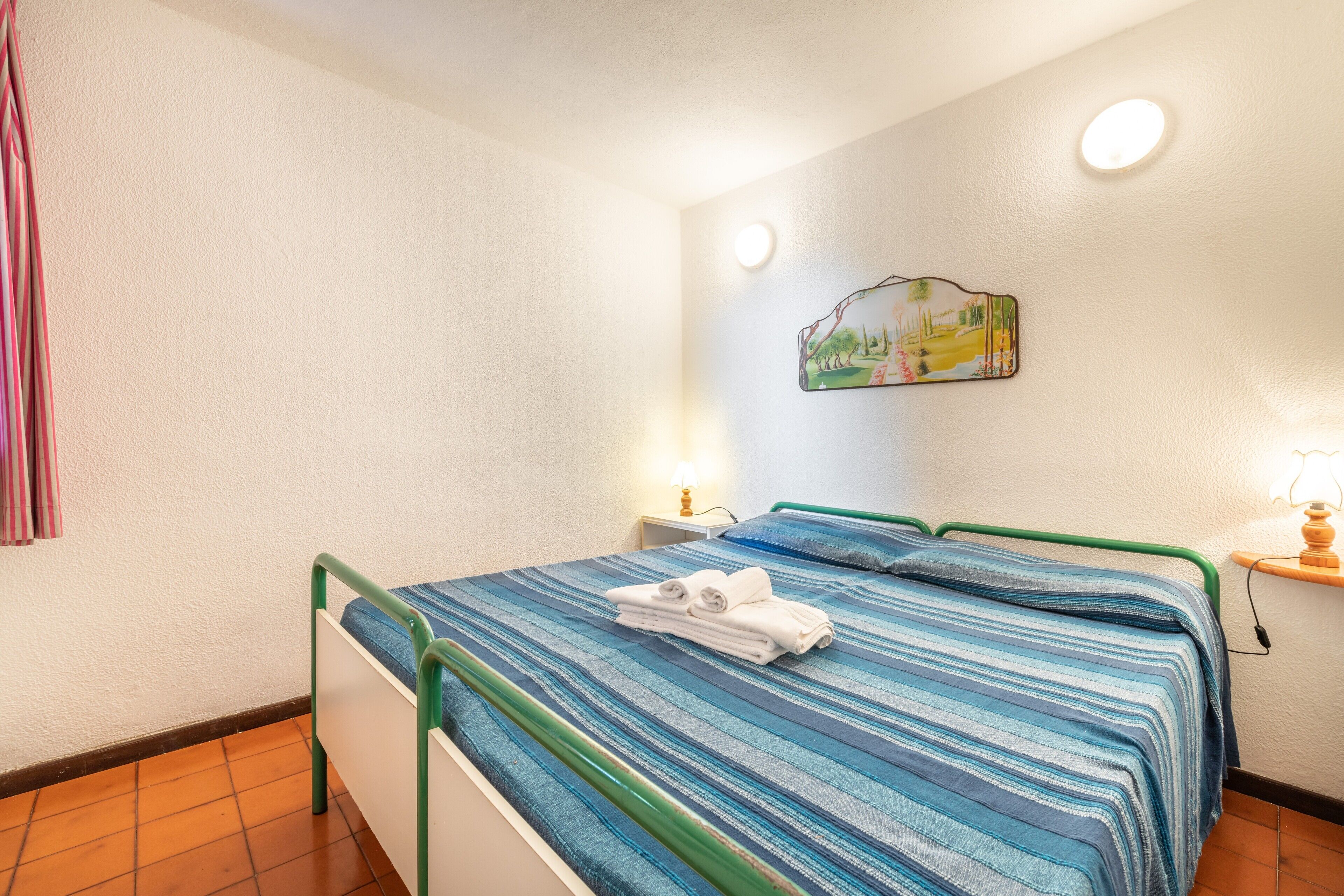A bedroom with a bed, a nightstand, a wall-mounted lamp, and a painting.