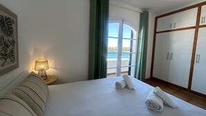 A hotel room with a bed, a nightstand with a lamp, a view of the sea through a window, and a wardrobe. - Beautiful Apartment with Wi-Fi, Pool, Terrace and Sea View (Arenal d'en Castell)
