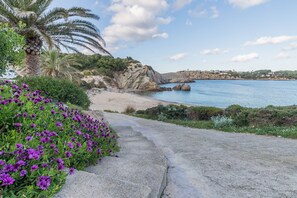 A coastal path with a stone walkway, leading to a beach with clear blue water, surrounded by greenery and a palm tree. - Beautiful Apartment with Wi-Fi, Pool, Terrace and Sea View (Arenal d'en Castell)