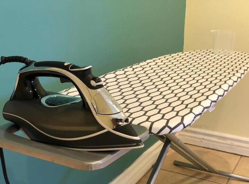 An ironing board with a patterned cloth, an iron, and a clear plastic cup on a table.