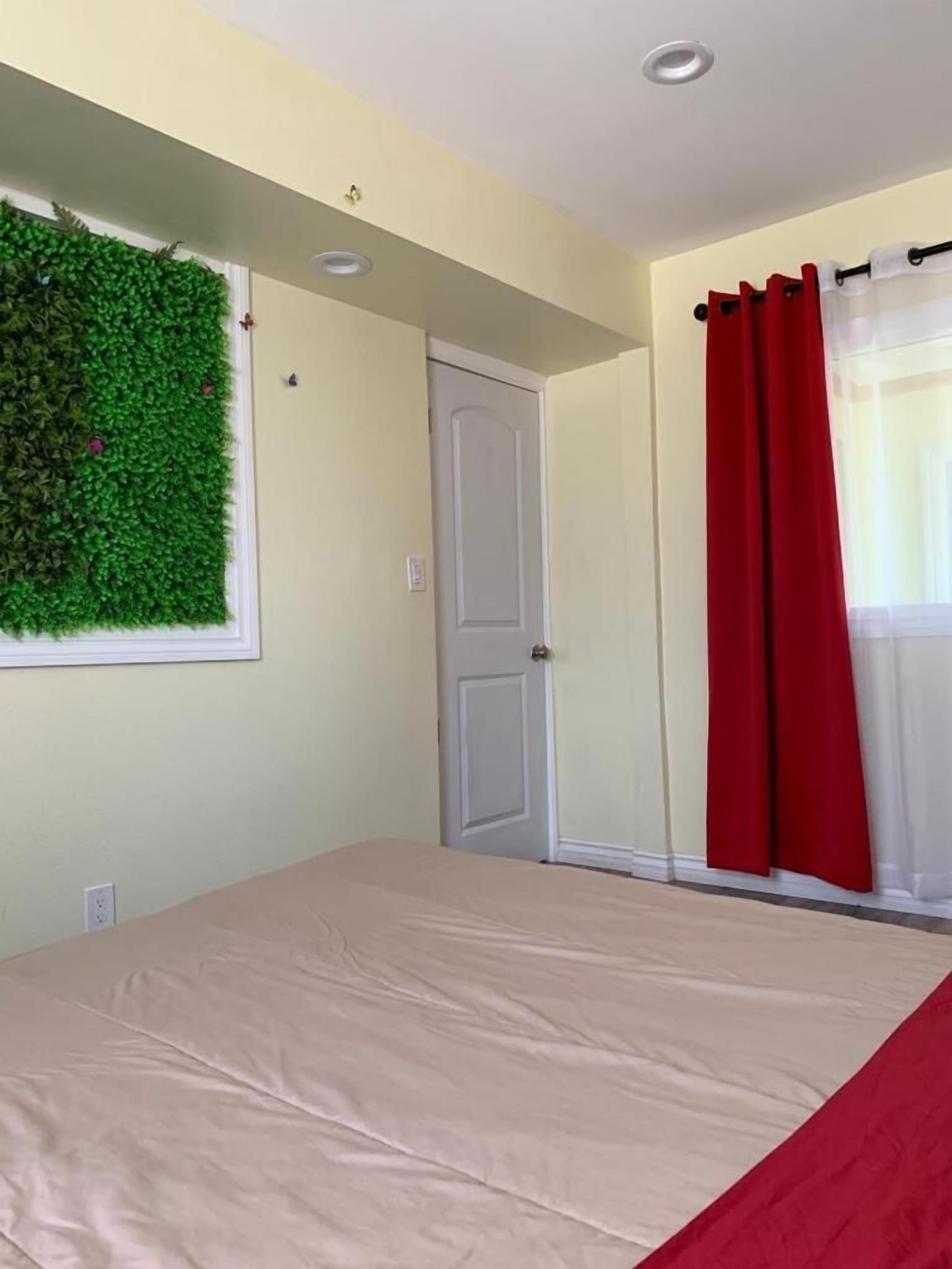 A bedroom with a bed, a door, a window with red curtains, and a green wall decoration.