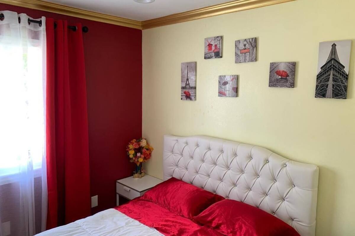 Un dormitorio con un cabecero blanco acolchado, una colcha blanca y roja, y una mesita de noche con un florero.