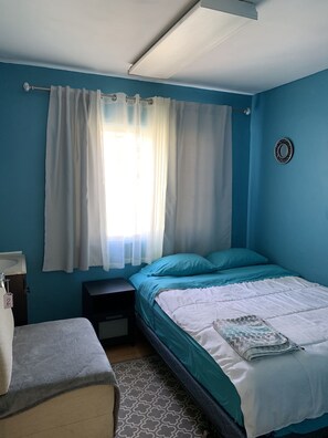 A bedroom with a bed, a nightstand, a small table, and a window with curtains. - LAX guest house (Inglewood)