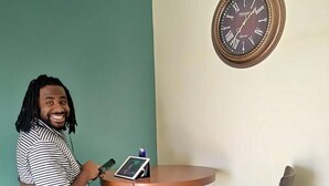 A person seated at a round table with a tablet and a can of drink, in a room with teal walls and a wall clock. - LAX guest house (Inglewood)