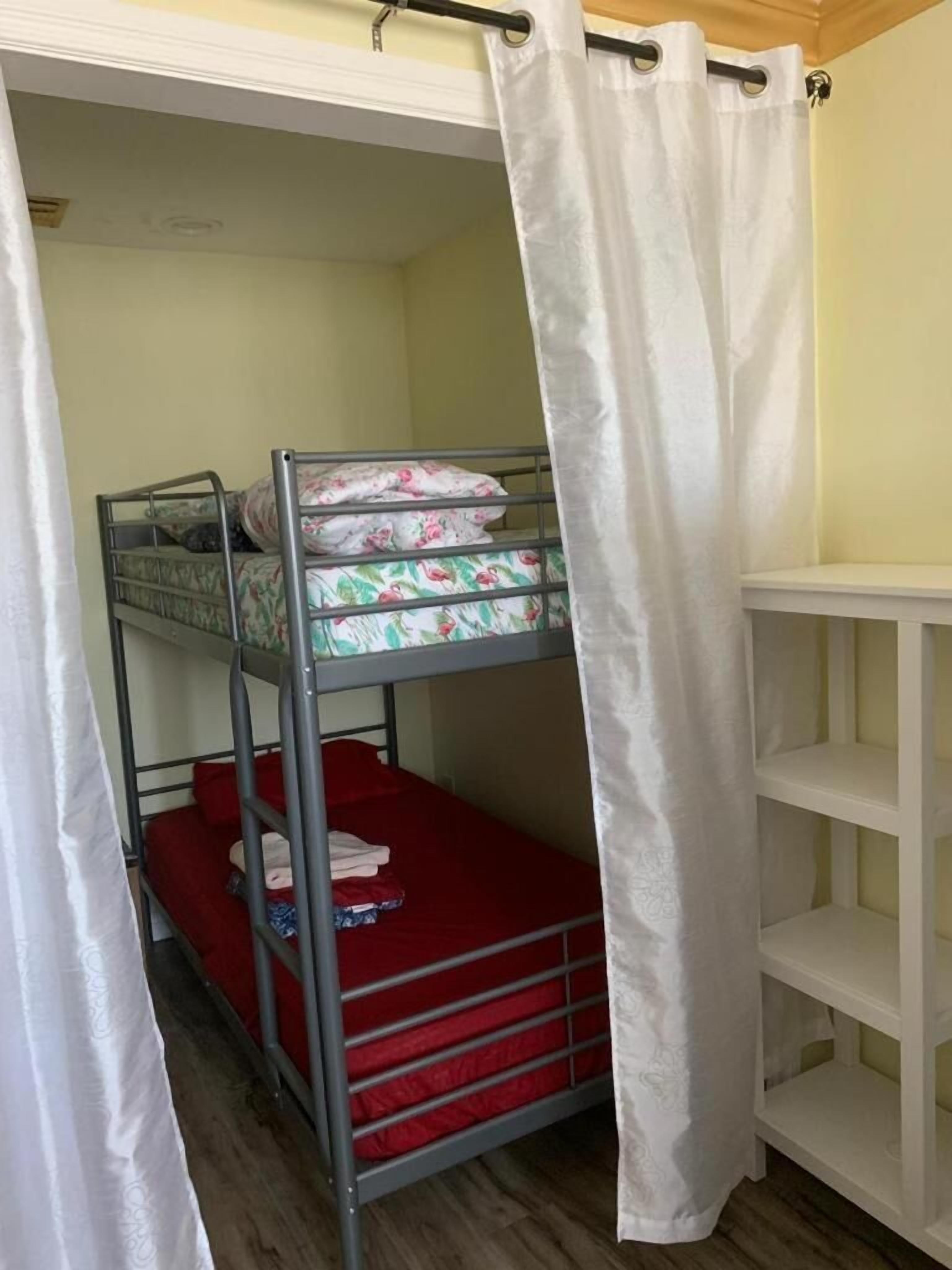 Dormitorio con literas, una cortina blanca, un colchón rojo y un estante blanco.