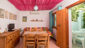 A room with a wooden table and chairs, a bed with colorful bedding, a television on a wooden cabinet, and a door leading to an outdoor area with plastic chairs.