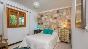 A bedroom with a bed, a dresser, a nightstand, a lamp, a window with curtains, and a stone-patterned wall.