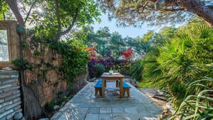 A stone pathway leading to a wooden table and benches, surrounded by lush greenery and a bamboo fence.