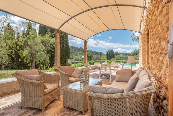 An outdoor patio with wicker furniture, a glass table, and a view of a pool and greenery.