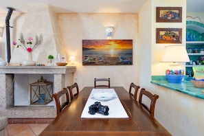 A dining area with a wooden table, chairs, a camera, a painting, and a kitchen counter with a lamp. - Beautiful Sun House Villa with Sea View, Wi-Fi, Balcony & Terrace (Cannigione)