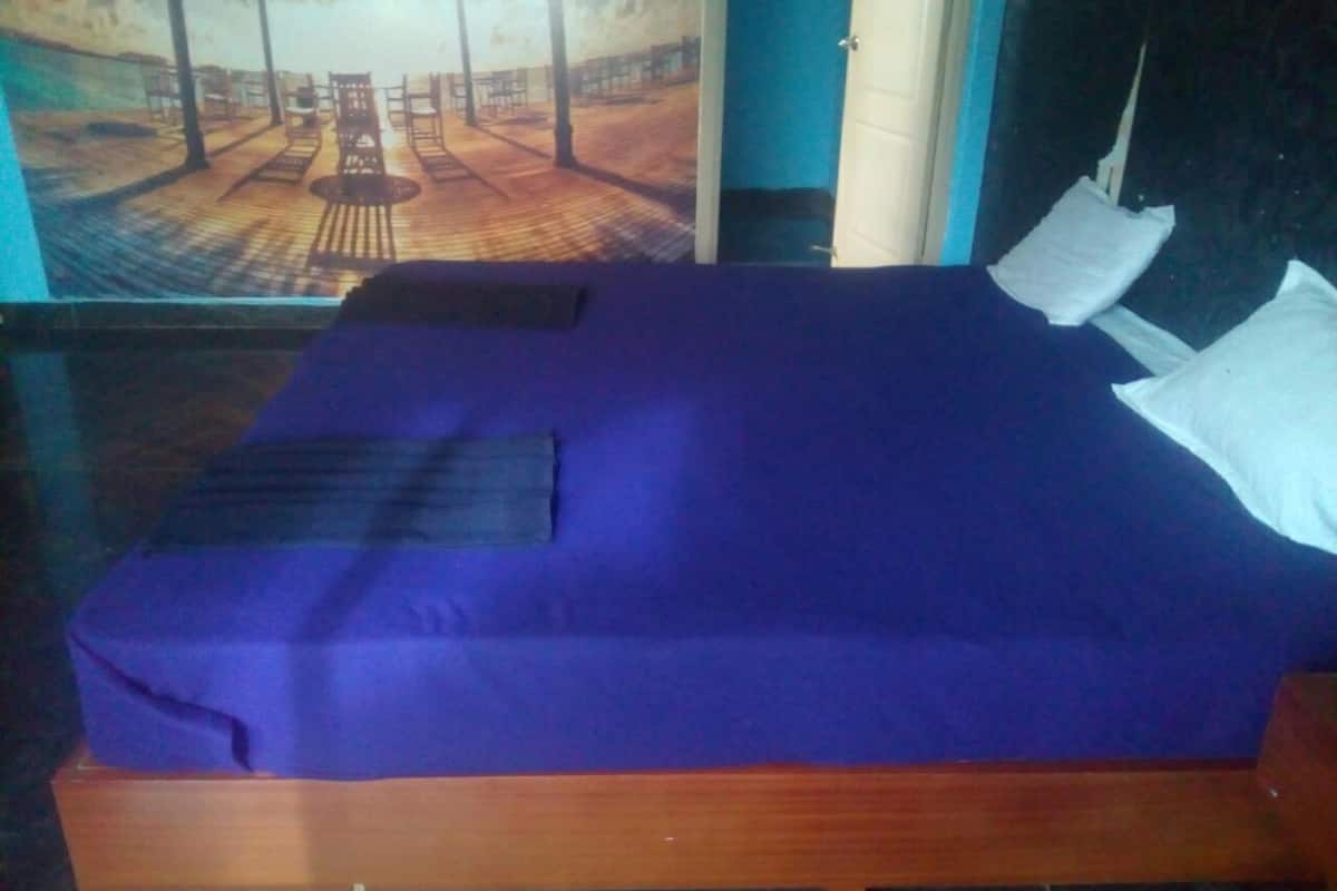 Een bed met een blauwe matras en witte kussens, een houten hoofdbord en een schilderij van een zandstrand aan de muur.